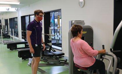 Woman using lat pulldown machine under supervision of man in UQ shirt